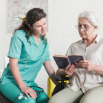 Disabled senior female patient sitting on wheel chair reading book with nurse
