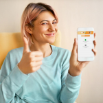 Charming happy young woman with pinkish hair holding smart phone with blank black display with copy space for your advertising content, making thumbs up gesture as sign of appoval, winking at camera