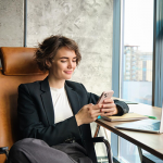 Portrait of businesswoman resting in her office in front of window using mobile phone sitting with